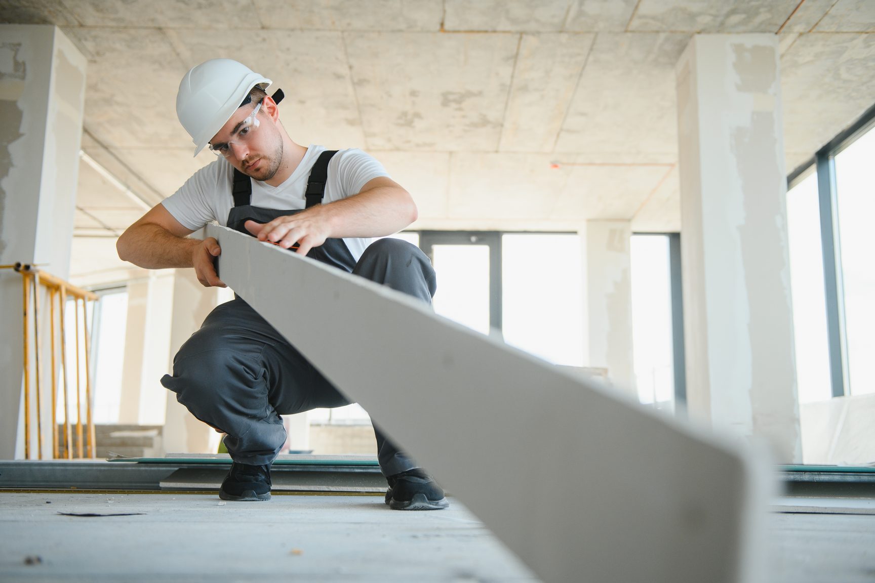 A worker in a white hard hat and safety glasses crouches on a concrete floor, handling a long sheet of drywall in an open commercial space.