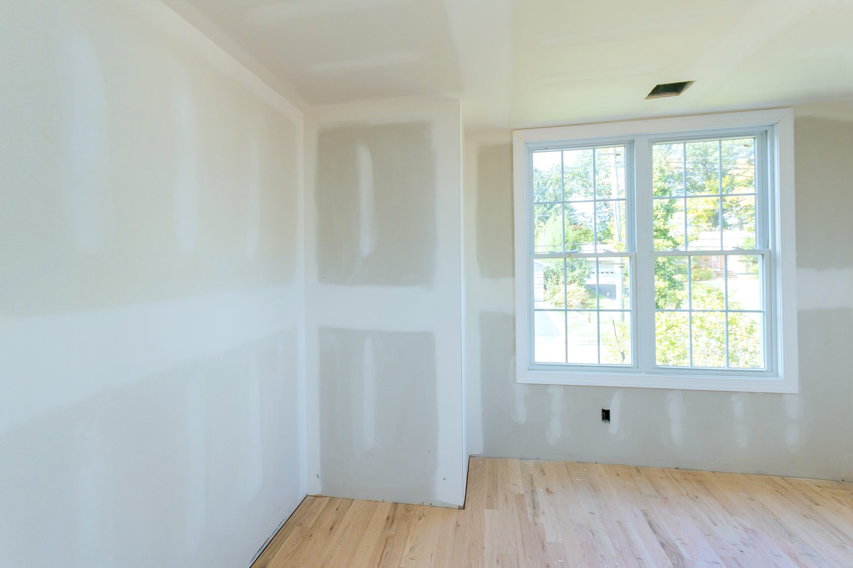 An empty room with taped and sanded drywall walls around a large window, a wood floor and openings in the ceiling for lights.