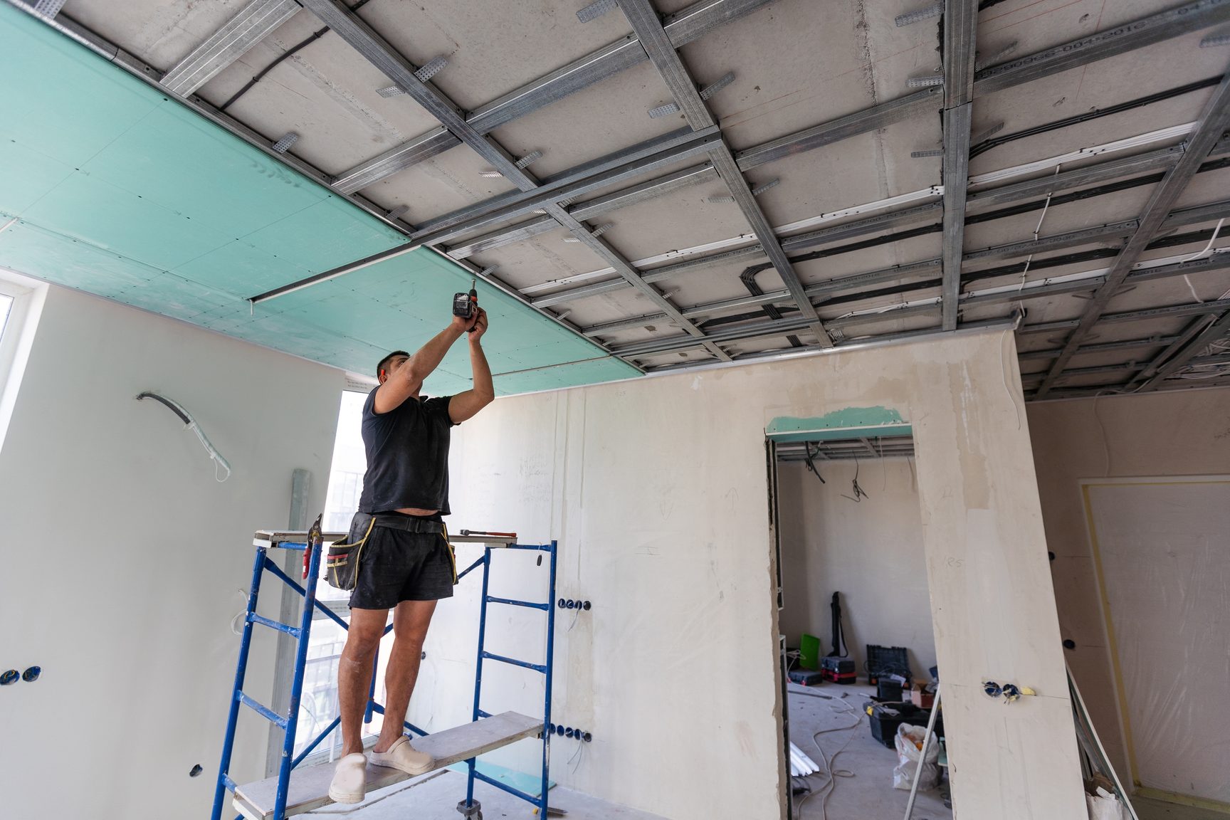 A worker on a rolling scaffold fastens drywall to a metal ceiling grid in an open commercial interior, with framed walls and a doorway below.