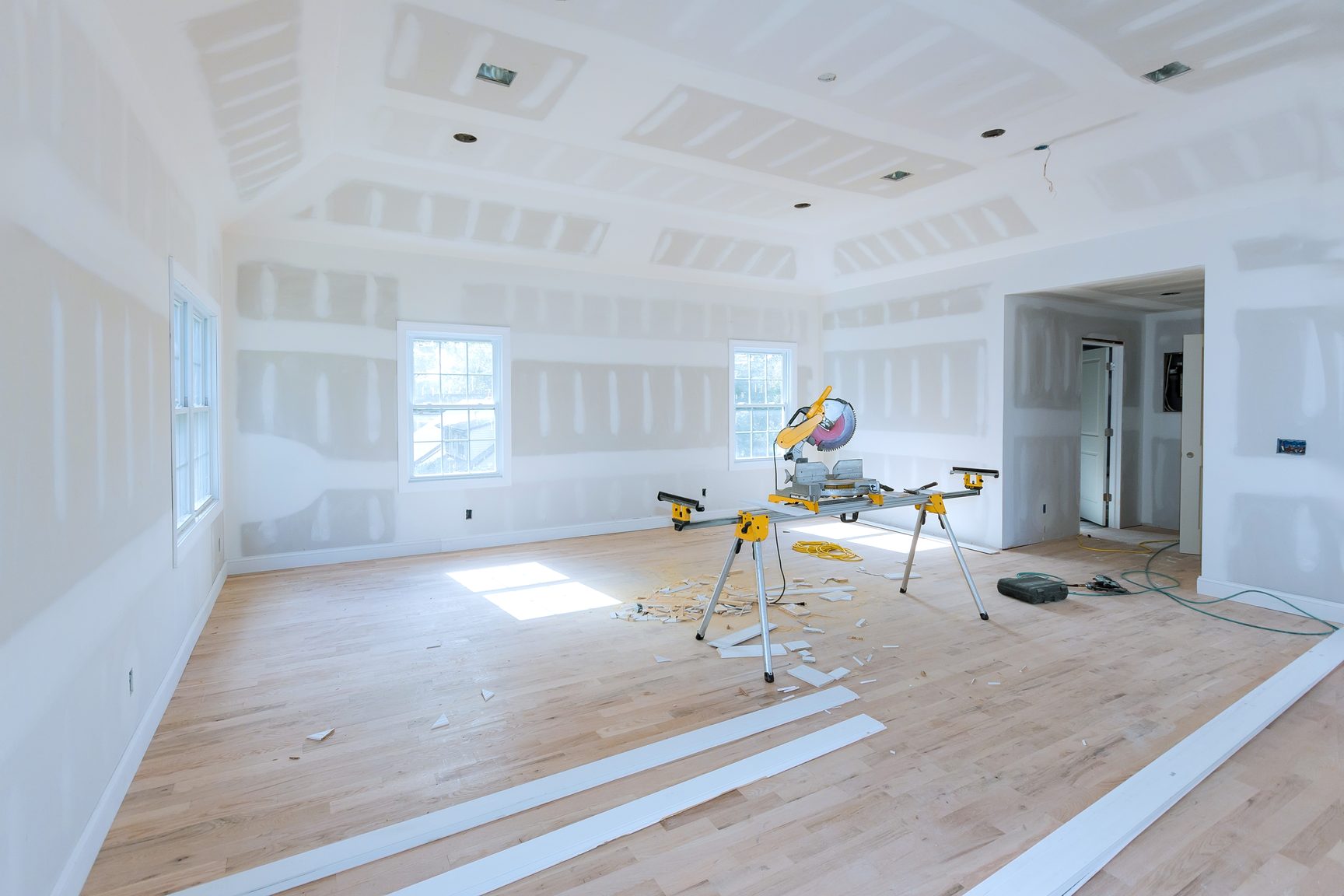 A large open room with freshly taped drywall walls and a coffered ceiling, a miter saw on a stand and trim stacked on a wood floor.