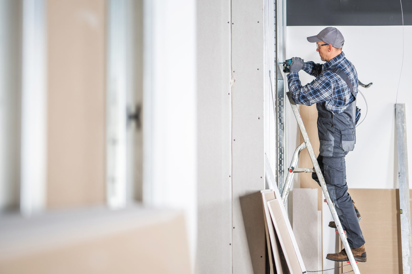 A worker in a cap and gray work clothes stands on a ladder fastening drywall beside a tall window in a bright interior under construction.